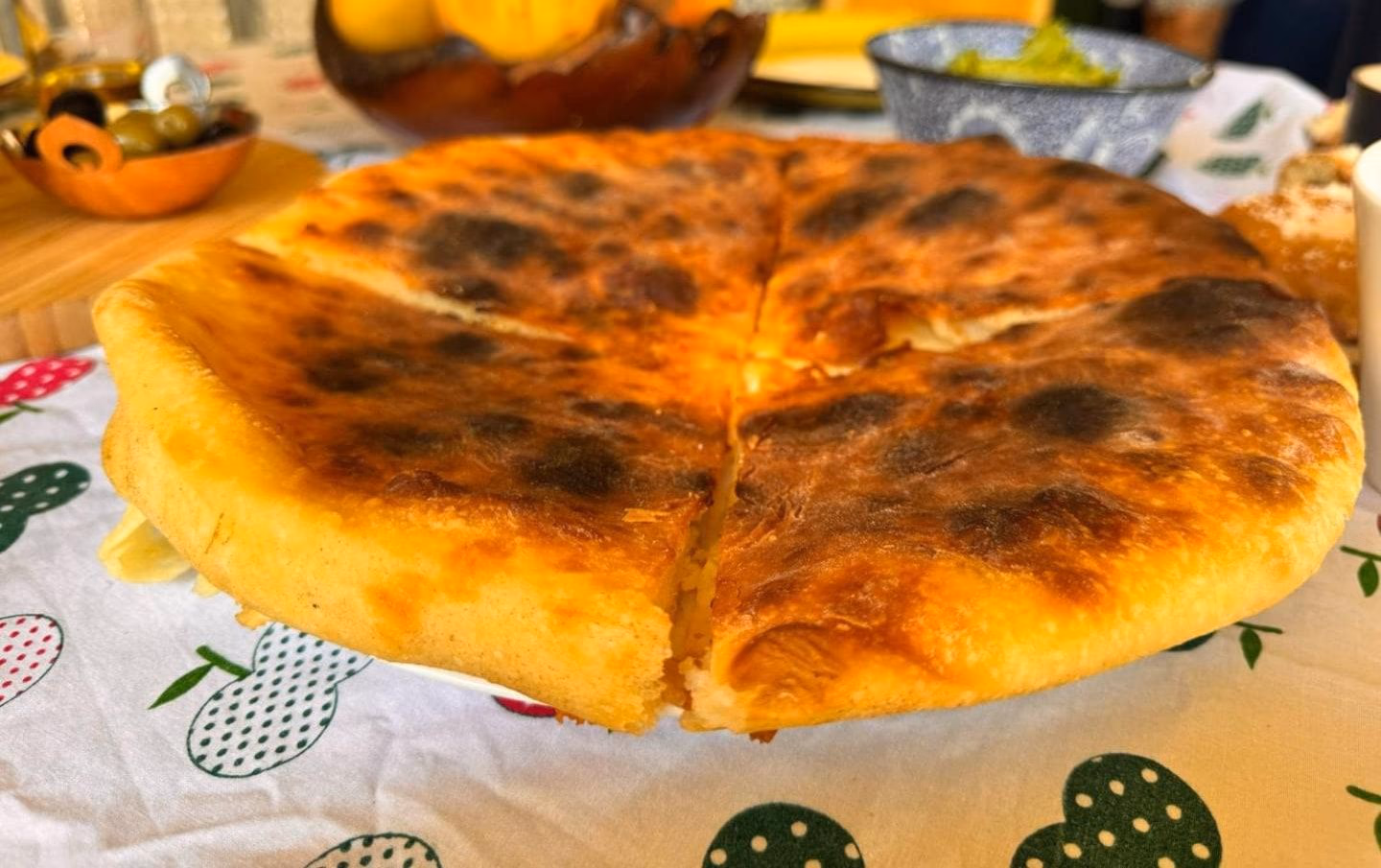 A whole imeruli khachapuri, quartered and blistered, on a fruit-print cloth with olives and fruit behind it