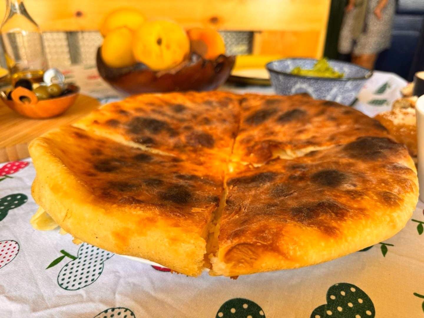 A whole imeruli khachapuri, freshly baked and blistered, on the table with peaches, olives and olive oil