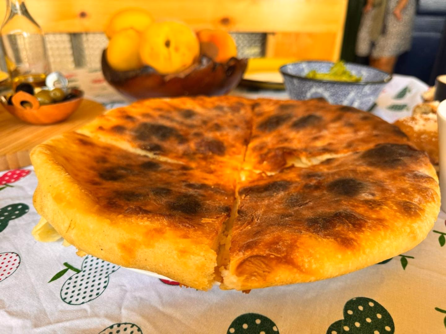 A whole imeruli khachapuri, blistered and golden, on the table beside peaches, olives and olive oil