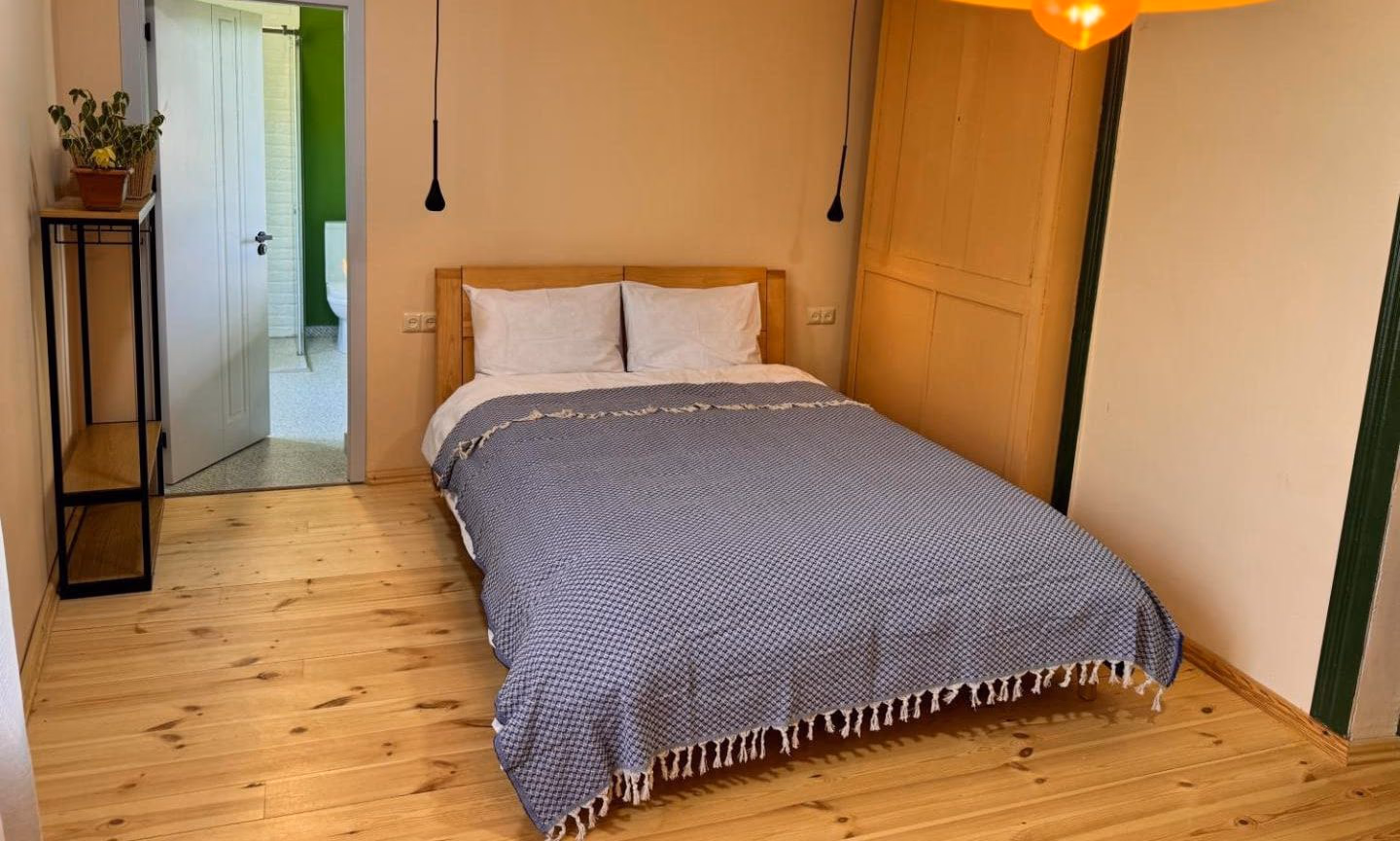 Bedroom with apricot walls, pine floorboards, an oak bed with a blue waffle throw, and a large olive-green dome lamp