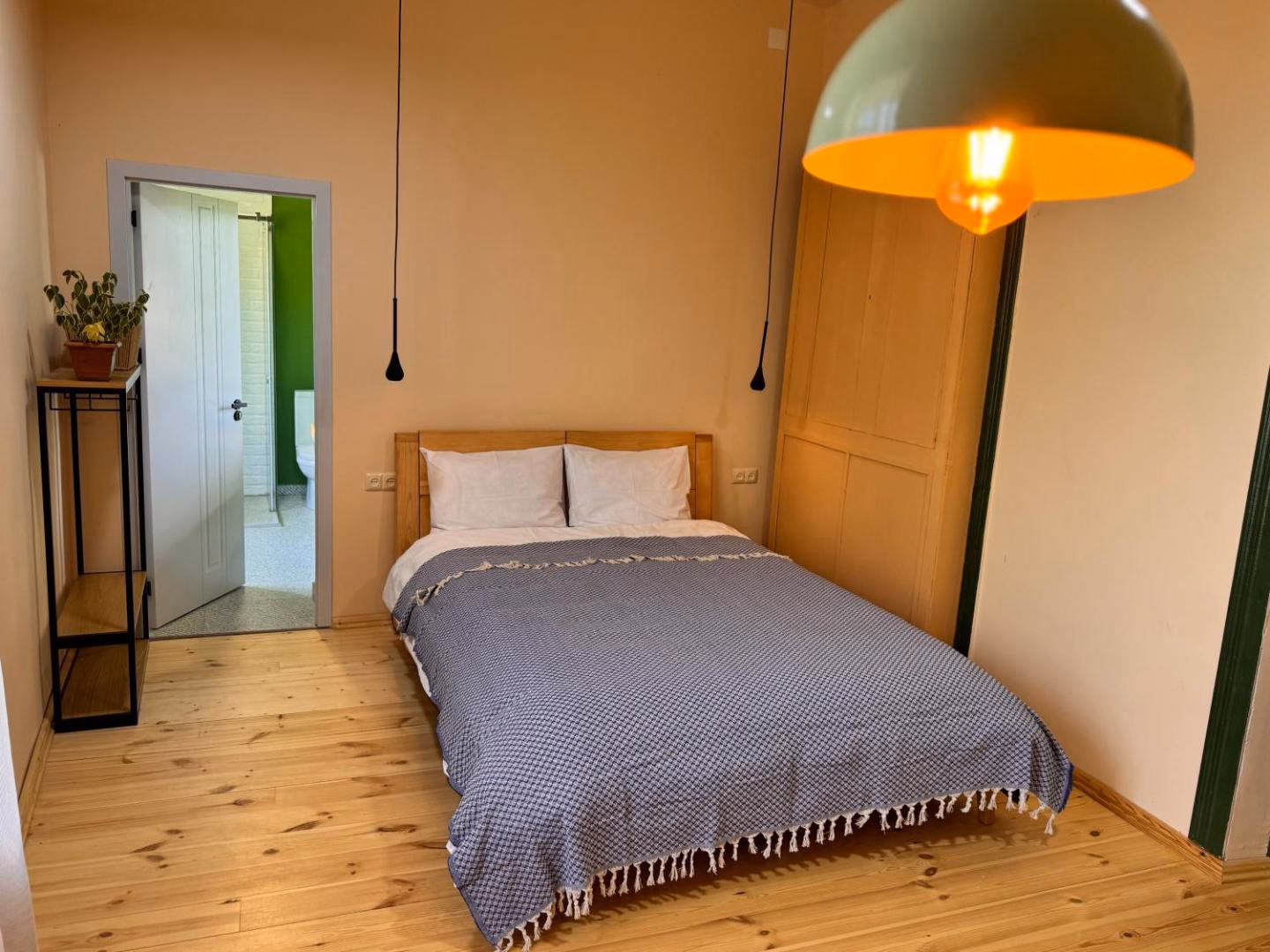 The Alazani room from the doorway: apricot walls, a black pendant over an oak double bed under a blue waffle cover, a mustard wardrobe, and the bright green bathroom through the open door