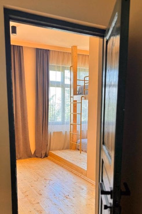 Looking through a dark green door into a room with pale pine floors, long curtains and a wooden bunk bed with a brass ladder beside the window