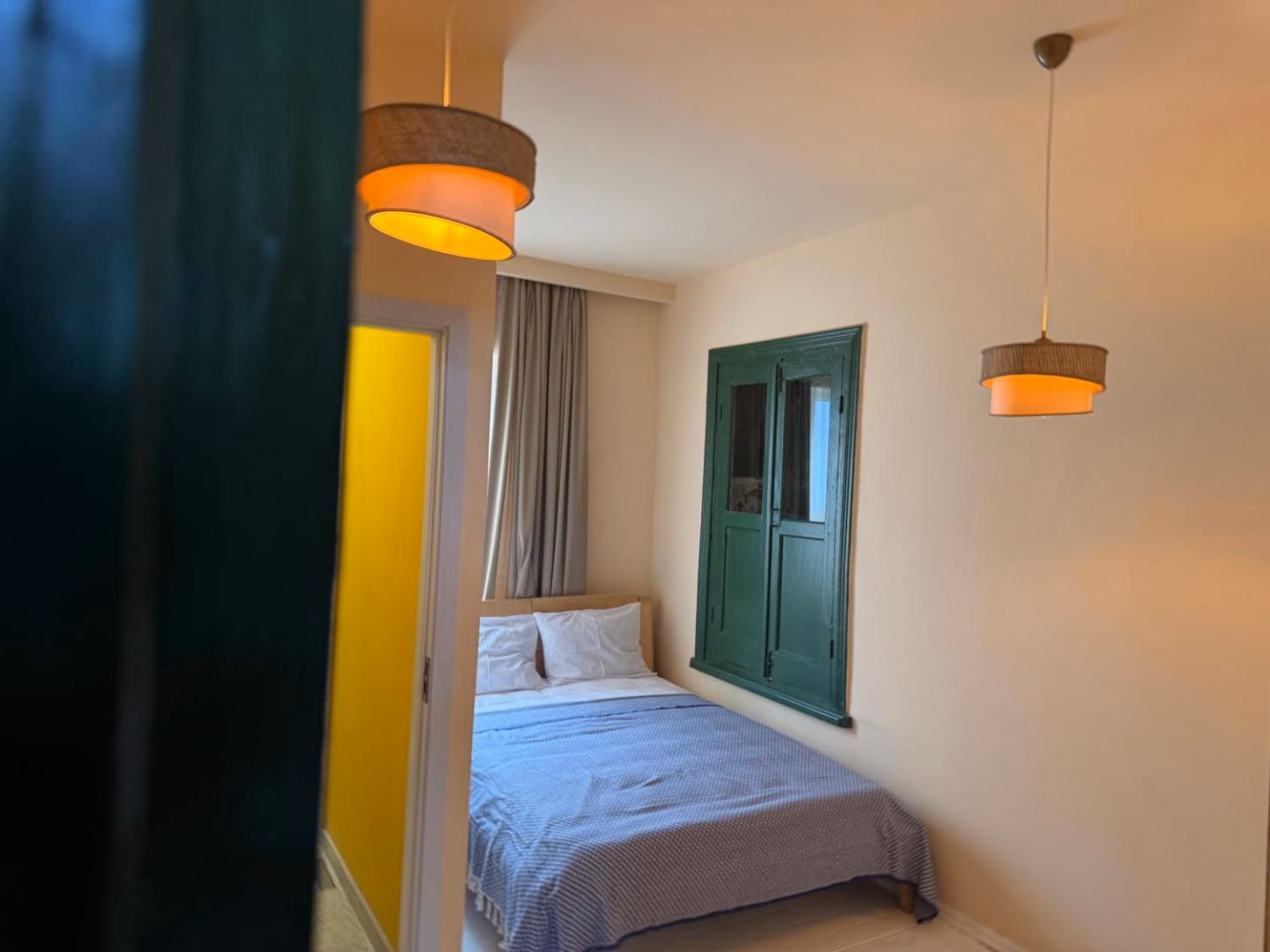 Marani from the corridor, past the edge of a dark green door: a small bed under a pale blue cover, the original green shutters set into the wall, and two woven pendant lamps