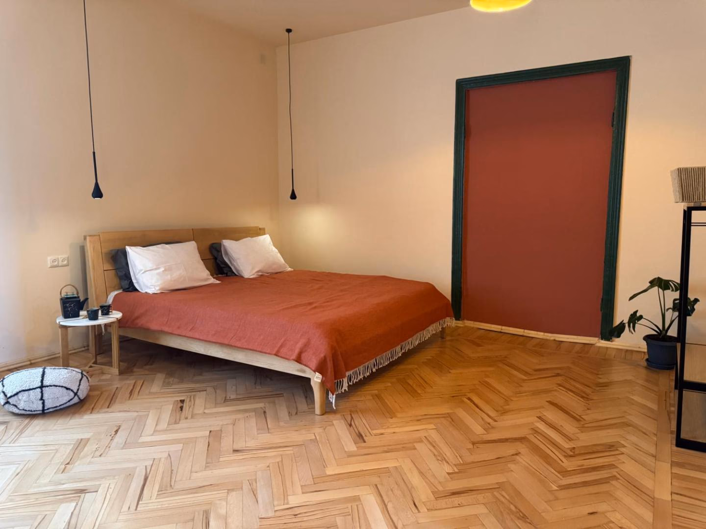 Large bedroom with herringbone parquet, an oak bed with a rust-coloured cover, and a terracotta door framed in green