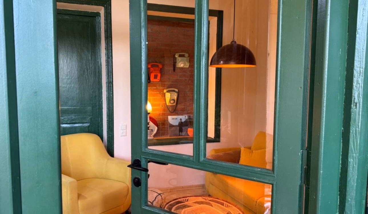 A green glazed door standing open between the hall and the sitting room, with a yellow armchair to the left and an exposed brick wall hung with two old telephones beyond it